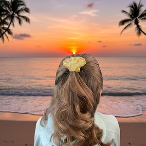 May include: A gold-colored seashell hair clip secures a ponytail. The person is facing away from the camera, with a beach sunset in the background. The sky transitions from orange to purple, with palm trees silhouetted on either side.