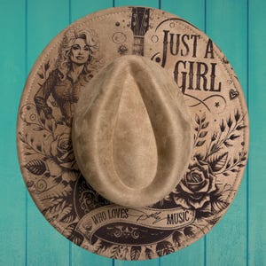 May include: A tan felt hat featuring a black printed design of a woman, a guitar neck, roses, and the text "JUST A GIRL WHO LOVES DOLLY MUSIC". The hat has a teardrop-shaped crown and a wide brim with a stitched edge, showcasing detailed line art.