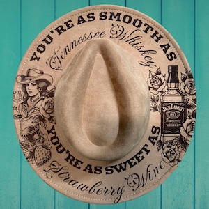 Könnte beinhalten: Beigefarbener Cowboyhut mit dem Text "YOU'RE AS SMOOTH AS Tennessee Whiskey" und "YOU'RE AS SWEET AS Strawberry Wine". Der Hut zeigt eine Grafik einer Frau, ein Hufeisen, eine Erdbeere und eine Jack Daniel's Flasche.