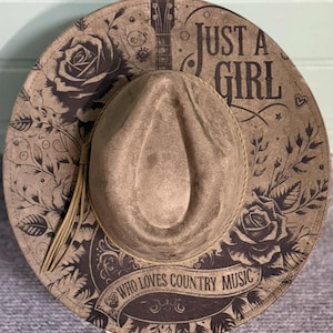 May include: A tan-colored fedora-style hat with a wide brim. The hat features a floral and guitar design with the text "Just a Girl Who Loves Country Music". A tassel hangs from the side.