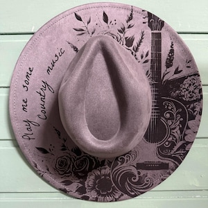 Puede incluir: Un sombrero de estilo fedora de color malva con un diseño floral y de guitarra decorativo. El sombrero presenta el texto "Play me some Country music" en una fuente manuscrita. El sombrero es un accesorio elegante.