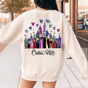 以下が含まれることがあります： クリーム色のスウェットシャツ。ディズニーの悪役が城の前にいて、ミッキーマウスの風船が描かれたカラフルなグラフィック。テキスト「Chillin' Like a Villain」が下にあります。