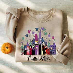 以下が含まれることがあります： ディズニーの悪役が城の前にいるグラフィックが特徴のベージュのスウェットシャツ。ミッキーマウスの風船付き。「Chillin' Like a Villain」の文字が下部にあります。左側には小さなオレンジ色のカボチャがあります。