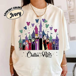 以下が含まれることがあります： アイボリー色のTシャツ。お城とディズニーヴィランズのグラフィックが特徴です。デザインには、ミッキーマウスの頭の形をした紫、緑、ピンクの風船が含まれています。Tシャツのテキストは「Chillin like a Villain」と書かれています。