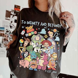 May include: Dark gray t-shirt featuring a colorful graphic of Toy Story characters, including Woody, Buzz Lightyear, and Jessie. The text "To infinity and beyond!" and "Toy Story Land" are displayed above the characters. The shirt also has "EST. 1995" printed at the bottom.