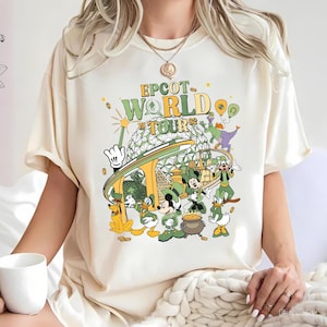 Puede incluir: Camiseta color crema con un gráfico colorido de personajes de Disney, incluyendo Mickey Mouse, Donald Duck y Pluto, con el texto "EPCOT WORLD TOUR 82". El diseño presenta una combinación de colores verde y dorado.