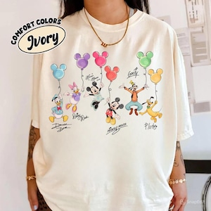 Könnte beinhalten: Elfenbeinfarbenes T-Shirt mit Disney-Figuren, die Mickey Mouse-förmige Ballons halten. Zu den Figuren gehören Donald Duck, Daisy Duck, Mickey Mouse, Minnie Mouse, Goofy und Pluto. Das Shirt trägt die Aufschrift "Comfort Colors" und "Ivory".
