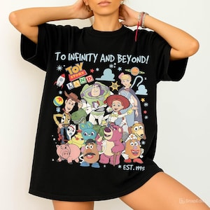 May include: Black t-shirt featuring a colorful graphic of Toy Story characters, including Woody, Buzz Lightyear, and Jessie. The shirt has the text "TO INFINITY AND BEYOND!" and "TOY STORY LAND" with "EST. 1995" below the characters.