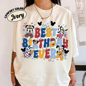 May include: Ivory-colored t-shirt with the words "BEST BIRTHDAY EVER" in colorful bubble letters. The design includes cartoon characters like Mickey Mouse, Minnie Mouse, Donald Duck, Goofy, and Pluto. The shirt has a Comfort Colors label.