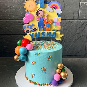 May include: A light blue frosted cake decorated for a first birthday. The cake is adorned with colorful sprinkles, edible balloon decorations, and a topper featuring a cartoon bus, characters, and the name "Yusha".