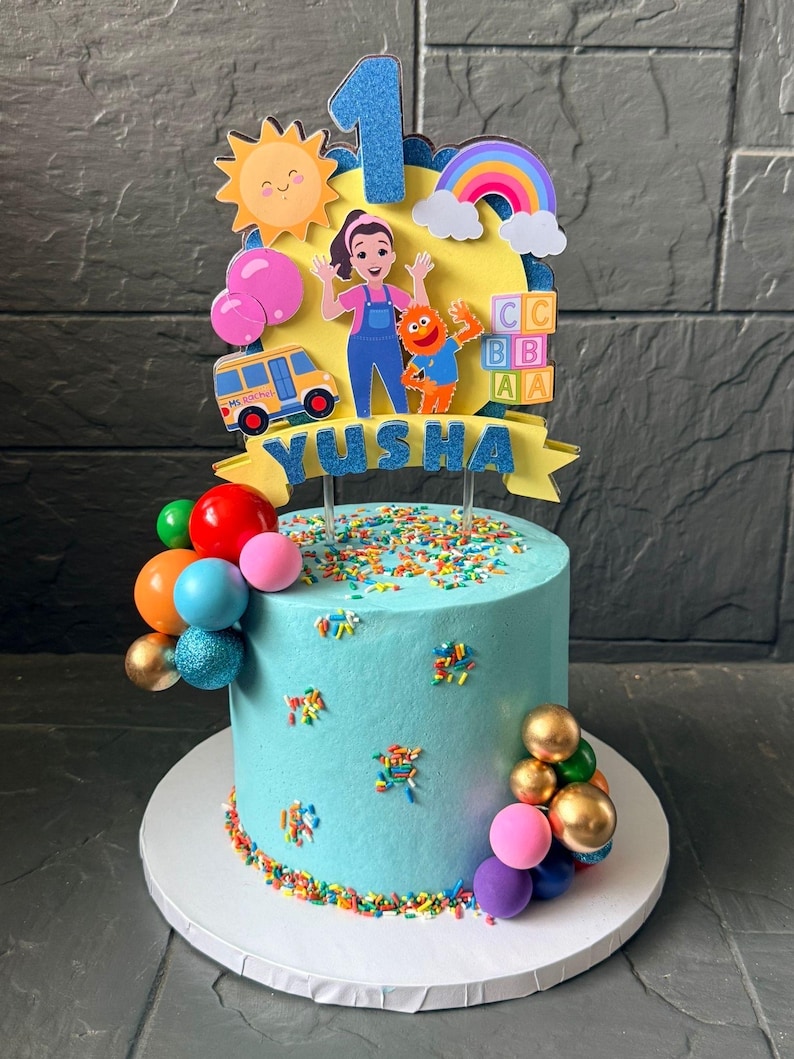 May include: A light blue frosted cake decorated for a first birthday. The cake is adorned with colorful sprinkles, edible balloon decorations, and a topper featuring a cartoon bus, characters, and the name "Yusha".