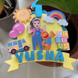 May include: A colorful birthday cake topper featuring a cartoon of a smiling sun, a rainbow, and a school bus. The topper includes the name "Yusha" in blue glitter letters and the number "1". The design also includes a cartoon of a woman and a monster.