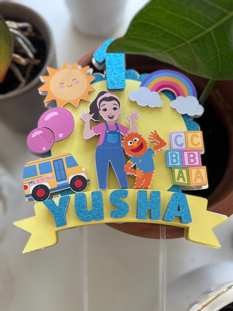 May include: A colorful birthday cake topper featuring a cartoon of a smiling sun, a rainbow, and a school bus. The topper includes the name "Yusha" in blue glitter letters and the number "1". The design also includes a cartoon of a woman and a monster.