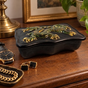 May include: A rectangular black box with a wavy edge featuring a raised, glossy green and gold dragon relief on the lid. The box has a matte finish and is positioned on a wooden surface alongside a wristwatch, gold cufflinks, and a gold chain.