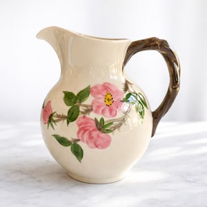 May include: Cream-colored ceramic pitcher with a floral design. The pitcher features pink flowers with yellow centers and green leaves. The handle is brown and has a textured, branch-like appearance. The pitcher has a spout for pouring.