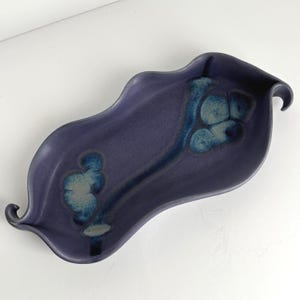 May include: An elongated, leaf-shaped ceramic tray with a matte deep purple finish. The surface features two distinct, organic-looking blue and white mottled glaze patterns that follow the central curve of the dish, which has curled ends.