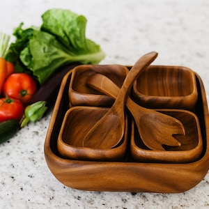 May include: A wooden salad bowl set with four square bowls and two salad serving utensils. The bowls and utensils are made of a warm-toned wood. Fresh vegetables, including tomatoes, carrots, and lettuce, are visible in the background.