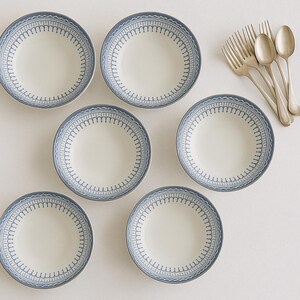 May include: A set of six white ceramic plates with a blue decorative border. Silver forks and spoons are arranged to the right of the plates. The plates have a simple, elegant design, suitable for everyday use or special occasions.