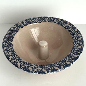 May include: A round ceramic baking mold with a smooth, light beige interior and a central pillar. The wide, flat rim features a dark blue and beige speckled pattern. The bowl has a glossy finish and a circular shape, designed for baking cakes.
