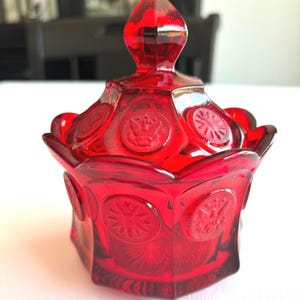May include: A ruby red glass candy dish with a lid. The dish is hexagonal with embossed starburst and medallion designs. The lid has a pointed finial and matching embossed medallions. The dish is likely vintage.