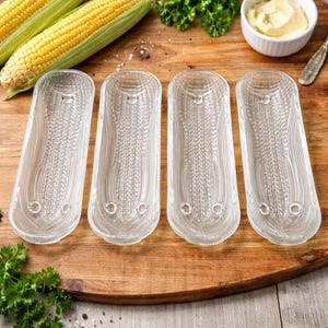 May include: Four clear glass corn-on-the-cob holders with a textured design, resembling corn kernels. The holders are arranged on a wooden cutting board, alongside fresh corn and a small bowl of butter.