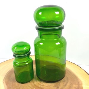 May include: Two translucent emerald green glass apothecary jars with rounded, dome-shaped lids. The jars feature a cylindrical body with a short neck and a wide rim. The glass is smooth and clear, allowing light to pass through the vibrant green color.