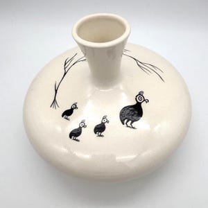 May include: Cream-colored ceramic vase with a rounded body and a cylindrical neck. The vase is decorated with black illustrations of birds and tree branches. The birds are stylized with large eyes and simple shapes. A unique decorative piece.