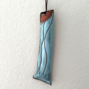 May include: A tall, slender ceramic wall vase featuring a light blue glazed finish with vertical wavy indentations. The top section is unglazed, revealing a reddish-brown clay texture with a carved architectural design of small building structures.
