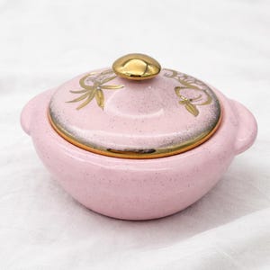May include: A pink ceramic lidded dish with a gold-colored knob and rim. The lid features gold floral designs. The dish has two small handles and a speckled texture. A decorative kitchenware item.