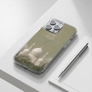 以下が含まれることがあります： モスクのデザインが施された透明な携帯電話ケース。ケースには、白いドームとミナレットを備えたモスクの詳細なイラストが、くすんだ緑色の背景に描かれています。ケースにはアラビア語のテキストも含まれています。