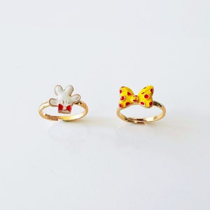 Puede incluir: Dos anillos de oro con diseños de esmalte de colores. Un anillo presenta una mano blanca con acentos rojos y el otro anillo presenta un lazo amarillo con lunares rojos.