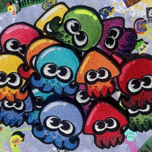 May include: A collection of embroidered patches featuring colorful squid and octopus characters with large white eyes and black pupils. The patches are rendered in vibrant shades of red, blue, green, orange, yellow, and purple with black outlines.