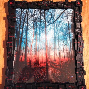 May include: A framed print featuring a forest scene with red and blue hues. The black frame has a geometric design with red accents. The artwork depicts tall trees with a vibrant, colorful landscape.