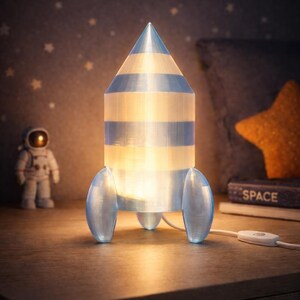 Op de afbeelding: Een raketvormige lamp met blauwe en witte strepen, van binnenuit verlicht, staat op een houten oppervlak. De lamp heeft drie vinnen en een puntige top. Een astronautenfiguur en een boek met het woord "SPACE" staan op de achtergrond.
