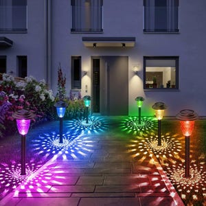 May include: A row of solar-powered garden lights with colorful light patterns illuminating a walkway. The lights have black poles and various colored glass tops, including purple, blue, green, yellow, and red. The lights cast a circular pattern on the ground.