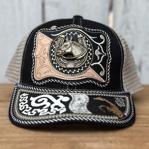 May include: Black trucker hat with a detailed horse head emblem. The hat features a mesh back, a curved brim, and decorative stitching. The front panel showcases a metallic horse head design with a horseshoe.