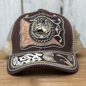 May include: Brown trucker hat with a decorative horse head and horseshoe emblem. The hat features a mesh back, a curved brim, and detailed stitching. The front panel has a leather-like texture with a metallic horse head.