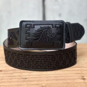May include: A black belt featuring a rectangular buckle with an embroidered horse head profile surrounded by a geometric border. The belt strap displays a repeating woven-style pattern in black thread, creating a textured appearance throughout.