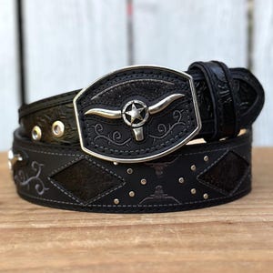May include: Black leather belt with a silver buckle featuring a steer head and star design. The belt has decorative stitching, silver studs, and a textured pattern. A stylish accessory for a Western-inspired look.