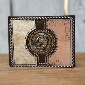 May include: A bi-fold wallet with a brown leather exterior, featuring a central design with a horse head emblem. The wallet has a light-colored, textured panel and a pinkish-tan panel, both framed with decorative stitching.