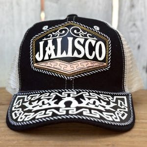 May include: Black trucker hat with "JALISCO" embroidered in white on a patch. The patch has gold and pink accents. The brim features a white geometric design. The hat has a white mesh back.