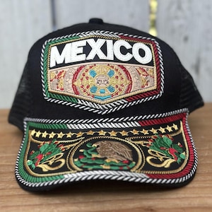 May include: Black trucker hat with embroidered "MEXICO" text and a detailed emblem. The emblem features a central design with red, green, and gold accents, and the brim showcases additional embroidery with the Mexican coat of arms.