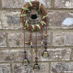 May include: A circular grapevine wreath adorned with green leaves and twine, featuring a central graphic of a cauldron and pentagram with the text UNSEEN ALCHEMY. Three dark red cords hang from the wreath, each holding small metal bells and glass vials.