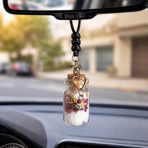 May include: A small glass bottle car charm filled with dried flowers and crystals, suspended from a black cord. The charm features a cork stopper and a decorative metal heart and star charm. The car's rearview mirror is visible in the background.