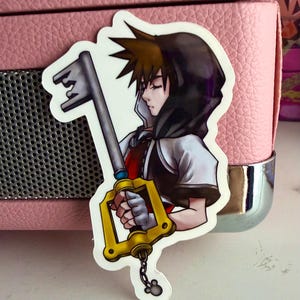 May include: A sticker featuring a character from the Kingdom Hearts video game series. The character is depicted with spiky brown hair, a black hooded cloak, and a key-shaped weapon. The sticker has a white border and is placed on a pink surface.