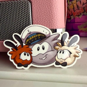 May include: A sticker featuring three cartoon characters. The central character is a gray creature wearing a sailor hat. Two smaller, fluffy creatures in orange and cream flank the central character. All three have black bow ties and smiling faces.