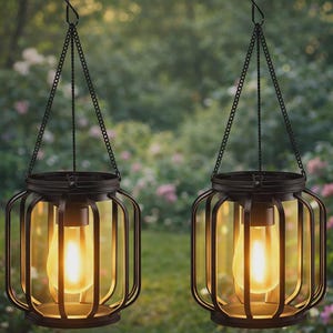 Könnte beinhalten: Zwei hängende Laternen aus schwarzem Metall mit warmem, leuchtendem Licht im Inneren. Die Laternen haben ein käfigartiges Design und sind an Ketten aufgehängt. Der Hintergrund ist eine verschwommene Ansicht von Grün und Blumen.