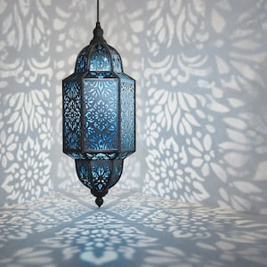 May include: A decorative hanging lantern with a dark metal frame and detailed cut-out patterns. The interior emits a bright blue light, creating floral shadows on the surrounding surface. The lantern's design is inspired by Middle Eastern aesthetics.