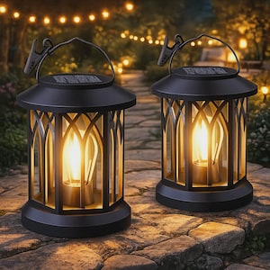 Puede incluir: Dos faroles solares negros con un diseño arqueado decorativo. Cada farol tiene un asa y una luz cálida y brillante en el interior, que se asemeja a una llama de vela. Los faroles están colocados sobre una superficie de piedra, con luces de cadena en el fondo.