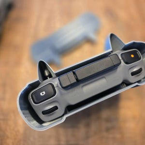 Puede incluir: Un mando de dron gris con botones negros y una funda protectora. El mando tiene un diseño elegante con un botón de encendido y otras funciones de control. La funda es de color gris claro y se ajusta perfectamente al mando.